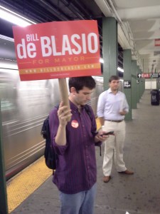 A de Blasio campaign staffer wields a sign, R train racing by. 