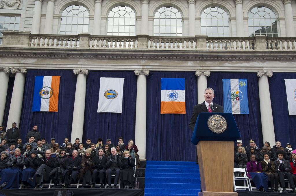 Mayor Bloomberg sitting expressionless in the background as Mayor Bill de Blasio speaks. (Official Photo: New York City Mayor's Office)
