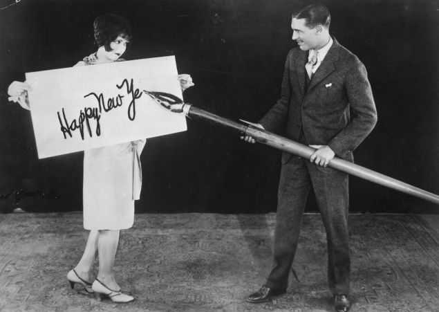 circa 1925: American actress Clara Bow (1905 - 1965) holds up a large card while actor Larry Gray inscribes a New Year's greeting with a giant pen. (Photo by General Photographic Agency/Getty Images)