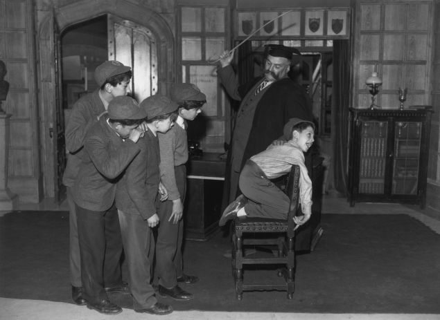 10th November 1959: 'Professor' Jimmy Edwards (1920 - 1988) in rehearsal at Shepherds Bush TV studios is administering a flogging to one of the pupils of 'Chiselbury' school, Paul Norman, watched by his classmates. (Photo by Reg Speller/Fox Photos/Getty Images)