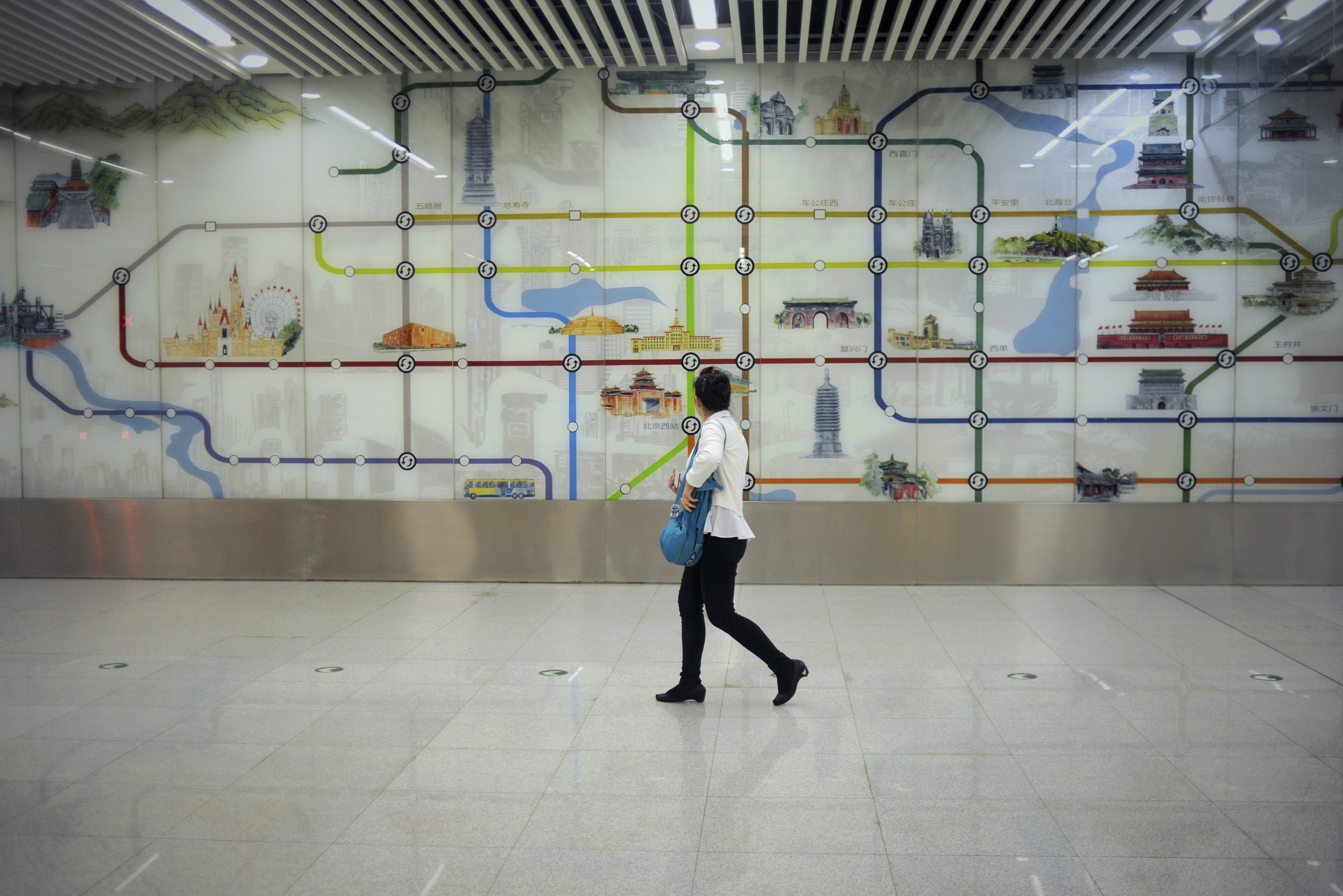 A woman walks past a wall painting of the Beijing subway system inside at a station in Beijing on May 9, 2016. As part of measures to try to limit the size of Beijing and relieve traffic congestion and reduce air pollution, some Beijing Subway lines or other commuter transport lines will expand to cities in Hebei. / AFP / WANG ZHAO