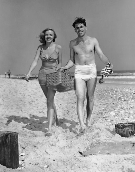 UNITED STATES - CIRCA 1950s: Couple walking with picnic basket on beach.