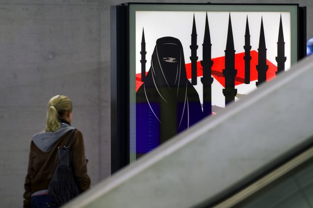 A woman passes by a campaign poster of the far-right Swiss People's Party depicting a woman wearing a burqa against a background of a Swiss flag upon which several minarets resembling missiles on October 26, 2009 at the central station in Zurich. Earlier this month, Switzerland's Commission Against Racism said that the anti-minaret poster campaign defamed the country's Muslim minority and could threaten public peace. Switzerland is expected to vote November 29 on a referendum launched by right-wing groups on whether the construction of minarets should be banned. AFP PHOTO / FABRICE COFFRINI 