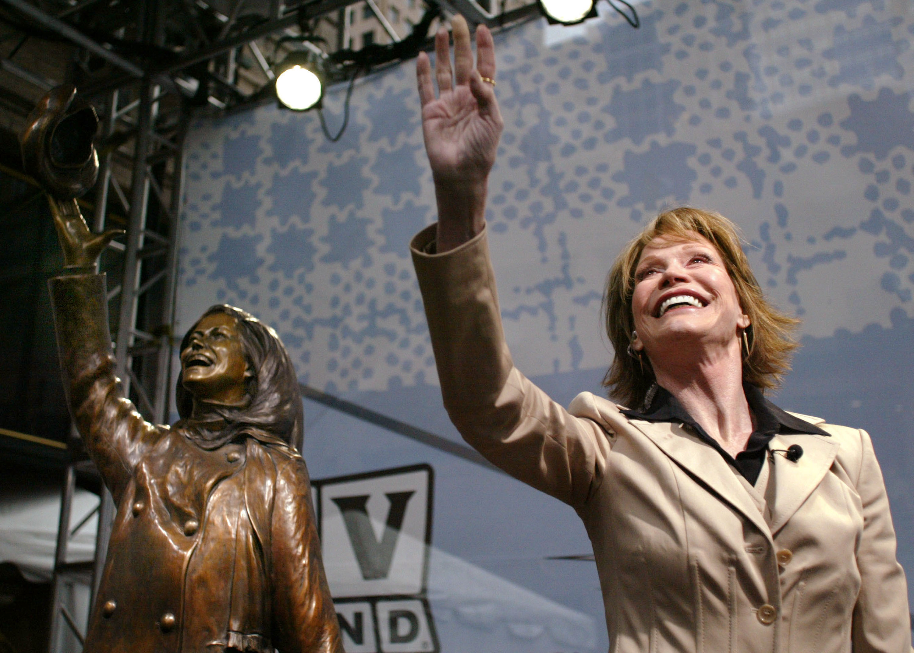 Mary Tyler Moore waves to the crowd next to a statue honoring her. The statue depicts Moore tossing her hat from the opening credits of The Mary Tyler Moore Show.