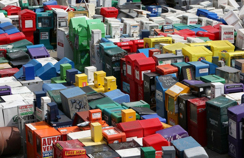 An assortment of newspaper and magazine vending racks sit in an empty lot.